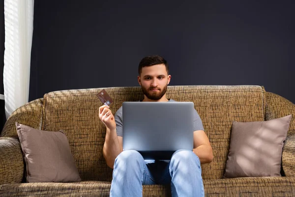 A young man in home clothes is sitting at home on a comfortable sofa with a laptop. Works and holds a bank card. Work online. Transfer of earned money. High quality photo