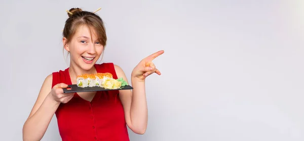 Banner- long format. Charming sexy caucasian woman with Japanese hairstyle appearance with sushi phyladelphia roll, pointing forefinger side on empty gray background. Space for text and publicity. — стокове фото