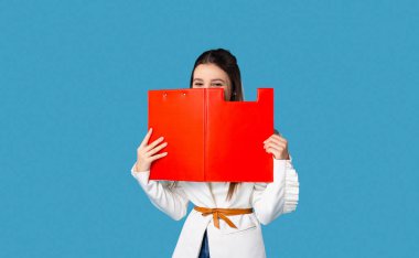 Sıradan giyinmiş, kırmızı panonun altında saklanan, mavi arka planda izole edilmiş boş bir alanın çekici ve hayalperest genç bayanın fotoğrafı..