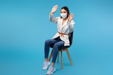 Koruyucu maskeli genç bir kız öğrenci mavi arka planda izole edilmiş bir sandalyeye oturur, iki eliyle hareket eder, bilgi almak için avuçlarını boş alana uzatır..