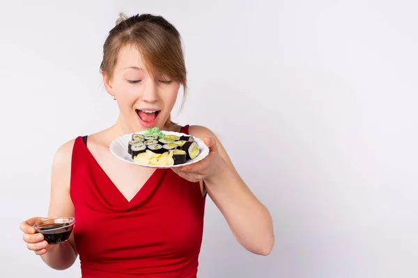 Стильна молода усміхнена дівчина позує, тримаючи тарілку рулетів і соєвий соус в іншій руці. Апетитна реклама для ресторану, суші-бар, доставка — стокове фото