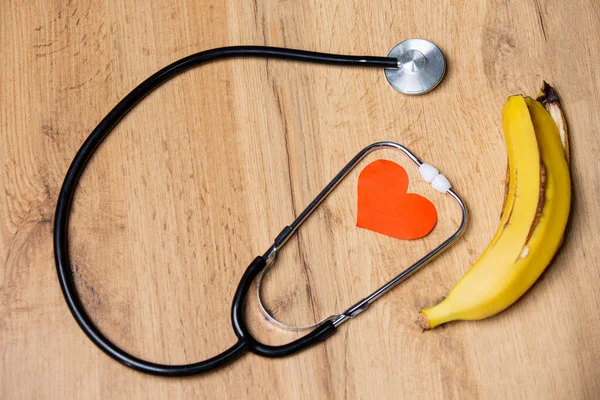 People with cardiovascular disease should consume potassium-rich bananas, which are antihypertensive food adjuvants. Paper heart stethoscope and banana on a table background.