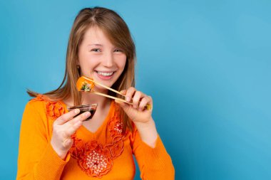 Sarışın, neşeli ve güzel bir kadının portresi. Yemek çubukları ve soya sosuyla suşi yiyor. Mavi arka planda izole edilmiş.