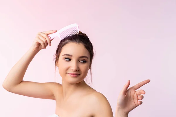 The femininity of a beautiful, cute brunette who comb her hair and points with finger to an empty spot. Young woman isolated on light pink background in beauty and care concept.