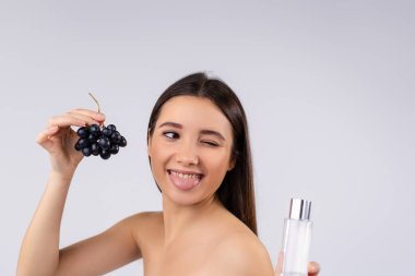 Taze üzüm ve bir bardak üzüm tohumu yağıyla esmer spa modeli. Yakışıklı kadın dilini gösteriyor, vücut ve cilt bakımı için kozmetik kullanıyor.