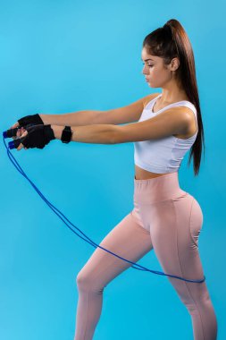 Esmer genç sporcunun profil görüntüsü mavi arka planda atlama halatıyla kardiyo egzersizleri yapıyor.