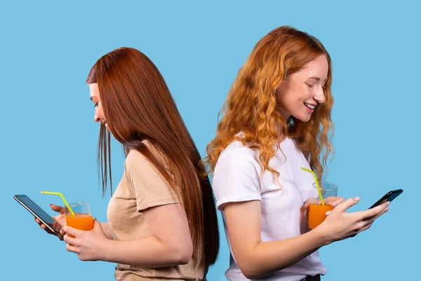 Two red-haired sisters back to back on a blue background with orange juice smile and spend a lot of time on social networks. Smartphone.