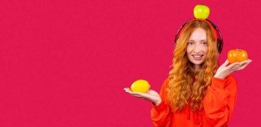 Banner, uzun format. Kızıl saçlı mutlu kız büyük kulaklıklarla müzik dinliyor ve kırmızı arka planda meyvelerle poz veriyor. Sağlıklı zaman geçirme kavramına sahip pankart.