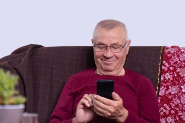 Gülümseyen yaşlı adam telefonu tutuyor, mobil cihaz uygulamaları kullanıyor, ekrana bakıyor, mutlu yetişkin erkek sohbeti yapıyor, mesaj yazıyor, alet koltukta otururken eğleniyor. Yüksek kalite fotoğraf