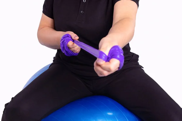 强烈的女运动员手与紫罗兰色的橡皮筋带坐在蓝色的健身球上 灰色背景的合影 高质量的照片 — 图库照片