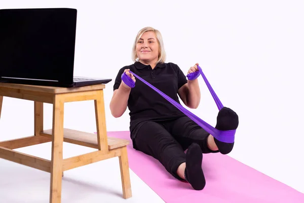 Menekşe fitness direnci elastik bantlı son sınıf bayan jimnastikçi trenleri dizüstü bilgisayardaki video bağlantısıyla eve dönüş, pembe yoga minderindeki uzaktan kumandalı sporlar. Yüksek kalite fotoğraf