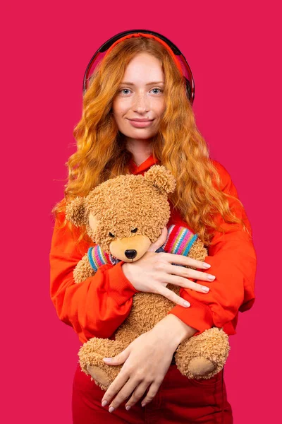 Healthy hair and perfect concept of freckled skin. Satisfied teenage girl with wavy red hair, headphones in her ears and a teddy bear in her hand, gentle smile, isolated over the red background. — Fotografia de Stock