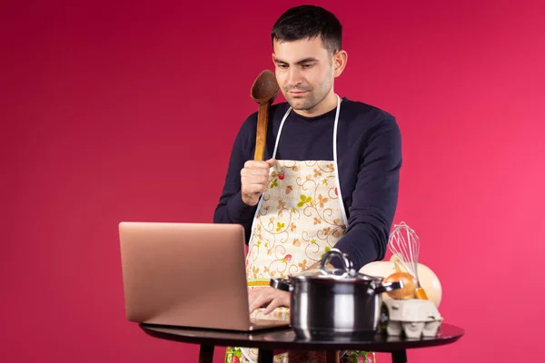 Online learning cooking class concept. Male stream videoblogger cooking and filming at the table in the kitchen. Cooking recipe modern blog using laptop. Stream blogger over red background — Fotografia de Stock