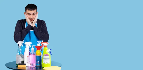 Banner over blue background, long format. Handsome man looking at all cleaning products, scared amazed, disbelief. Mature middle in cleaner apron covering face with hands, shocked afraid surprised — Fotografia de Stock