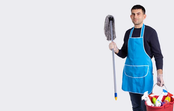 Health protection. Cleaning service concept. Young man wearing blue apron and rubber gloves with cleaning tools and products posing on gray background with empty side space for your ad — Stock Photo, Image