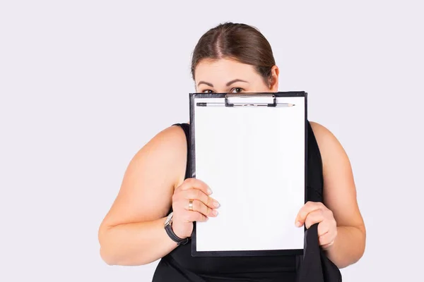 Closeup shot of mature businesswoman, plus size, overweight peeking over black clipboard with blank mock up white sheet with suspicious, distrustful face expression. Gray background — Foto Stock