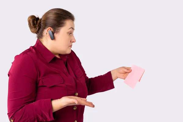 A new message alert. Plump girl, businesswoman wearing bordo shirt, talking with furious and doubtfull expression, wireless earphones, hands free, daily mail, pink blank envelope. High quality photo
