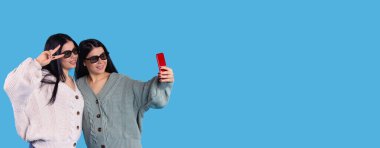 Banner, uzun format. Genç kadın ikizler 3 boyutlu gözlüklerle poz verirler ve akıllı telefondan selfie çekerler. Mavi stüdyo arka planında izole edilmiş, mesaj veya reklam için boş alan var. Yüksek kalite fotoğraf