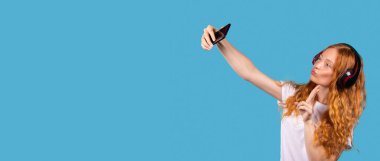 Afiş, uzun format, mavi arkaplan. Güzel beyaz kadın hava öpücüğü gönderir, barış jesti yapar, selfie çekmek için poz verir, akıllı telefon cihazına, stüdyo çekimine, kulaklıkla müzik dinleyerek mutlu olur.