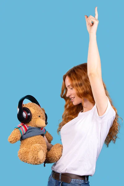 Κοκκινομάλλα νεαρό χαριτωμένο κορίτσι χορεύουν κοιτάζοντας αρκουδάκι της με ακουστικά ακούει μουσική. Μπλε φόντο, gadget, συσκευή, τεχνολογία έννοια με κενό χώρο για διαφήμιση — Φωτογραφία Αρχείου