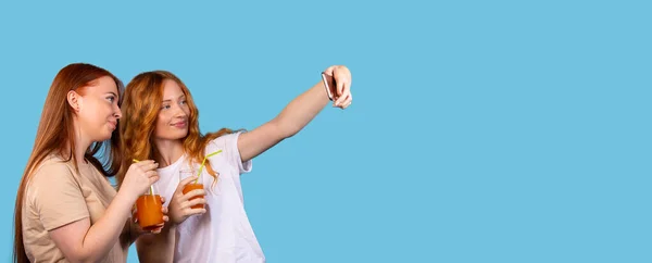 Banner, formato largo, fondo azul. Mujeres jóvenes felices en ropa casual centrándose en gadget de pantalla tomando selfie con smartphone sosteniendo gafas con bebida naranja. Espacio de ocio estilo de vida para el anuncio — Foto de Stock