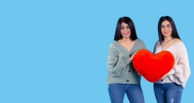 İki ikiz kız ellerinde pelüş bir kalp tutuyorlar ve kameranın önünde gülümsüyorlar. Mavi bir arkaplanda izole edilmiş