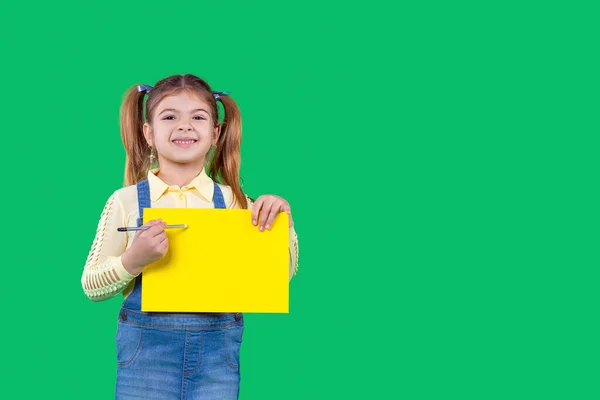 Una niña sostiene un lápiz en sus brazos y un papel de color amarillo. Lección educativa. Fondo verde — Foto de Stock