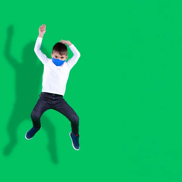 Niño en la forma de la escuela, máscara textil en el tiempo de descanso en la escuela. Foto de un chico saltando sobre un fondo verde. — Foto de Stock