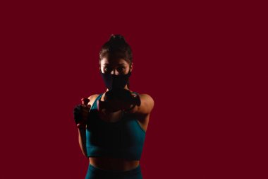 Spor eldiven giyen genç bir kadın, çok işlevli bir maskeyle boks yapıyor ve koyu kırmızı arka planda kameraya bakıyor. Yüksek kalite fotoğraf