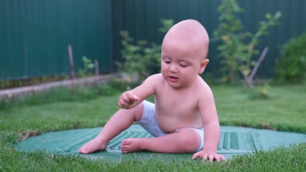 Bonito pequeno bebê solteiro em shorts está jogando ao ar livre com a grama verde, ele tenta arrancá-lo. — Vídeo de Stock