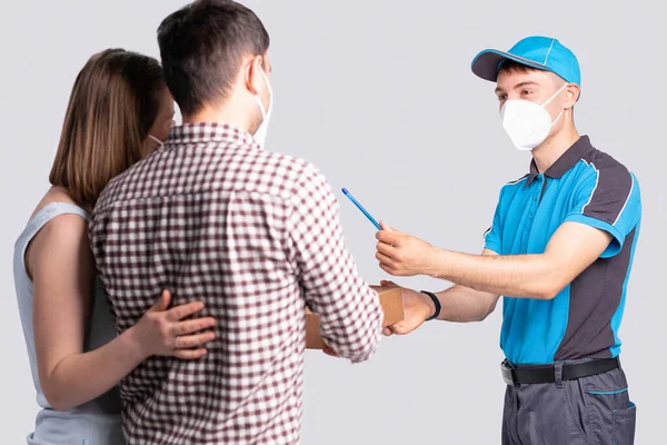 The courier in a mask is holding out a pen to confirm the delivery of the parcel for the couple in a mask. — Stock Photo, Image