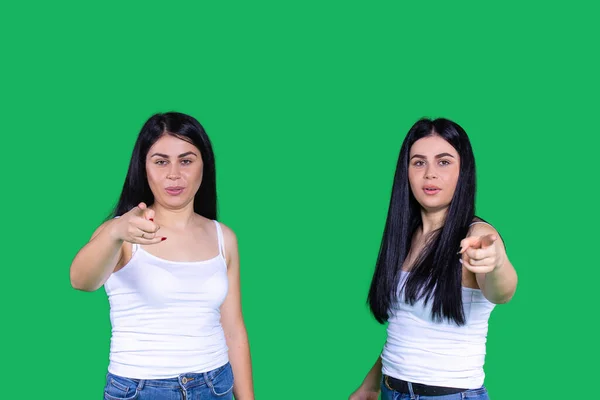 Хоробрі молоді дівчата-близнюки серйозно дивляться на камеру і показують свої індексні пальці на ній на зеленому тлі . — стокове фото