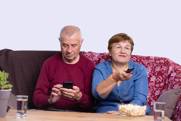 An old nice couple are resting sitting on the sofa, wife is switching channels on the remote and husband is working with cellphone. — Φωτογραφία Αρχείου