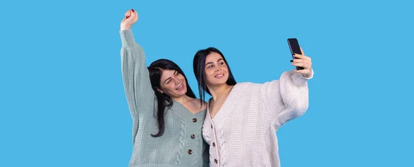 Mavi arka planda izole edilmiş selfie çeken neşeli ikiz kızlardan biri sağ elini kaldırıp gülümsedi.. — Stok fotoğraf