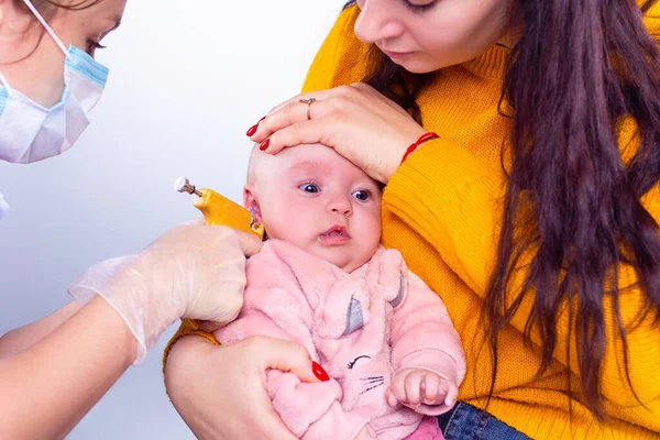 Atento mamá sosteniendo a su bebé por la cabeza y cosmetólogo con máscara médica y guantes perfora las orejas. — Foto de Stock