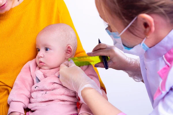 En el salón cosmético la mujer el cosmetólogo en la máscara miden con la regla el lugar para los pendientes para la chiquitina. — Foto de Stock