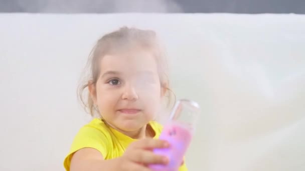 Маленькая девочка в жёлтой футболке сидит на диване, держа в руках увлажнитель и экспериментирует с ним прямо перед видеокамерой. — стоковое видео