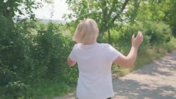 Ωραία ξανθιά μεσήλικη γυναίκα που γυμνάζεται τρέχοντας στο μονοπάτι ισιώνει τα μαλλιά και στρίβει προς την κάμερα κουνώντας το χέρι. — Αρχείο Βίντεο