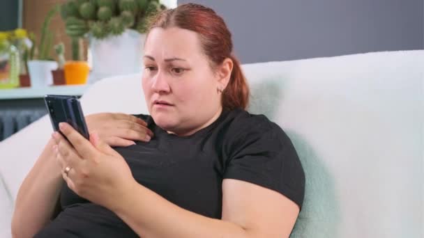 Mujer de gran tamaño sentada en el sofá de la sala de estar buscando malas noticias en el teléfono de Internet y sintiéndose con ella tener la expresión de la cara preocupado. — Vídeos de Stock