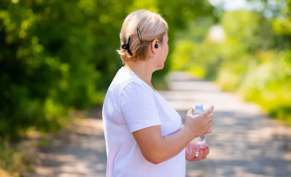 Photo latérale dos d'une femme blonde forte prend soin de sa santé jogging le matin tenant une bouteille d'eau et en utilisant un casque. — Photo