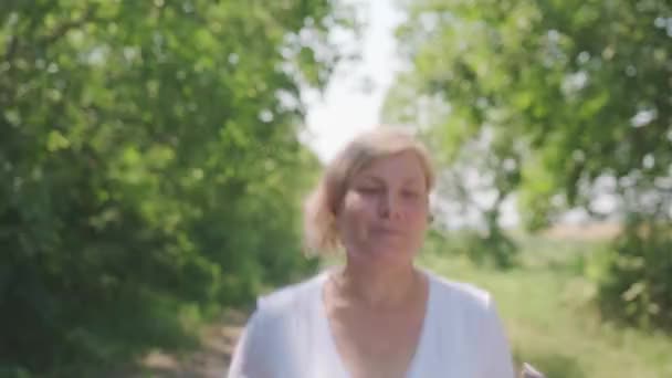 Das frontale Video einer lächelnden erwachsenen Frau, die im Freien joggt und dabei die Digitaluhr in der Hand hält, um sich in guter körperlicher Verfassung zu halten. — Stockvideo