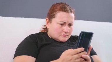Beyaz koltukta oturan bir kadın cep telefonunu kollarında tutuyor ve internette duygusal haberler seyrediyor..