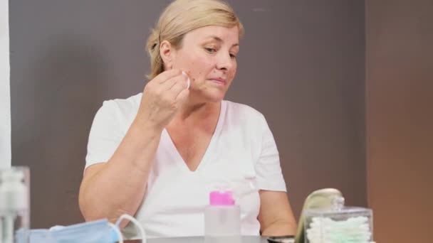 Smiling blonde beautiful woman sitting at the table trying to get rid of maskcne using the cotton disk blotting it with lotion and wipes her face. — Stock Video