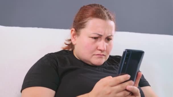Femme assise sur le canapé blanc tenant le téléphone portable dans ses bras et regardant des nouvelles émotionnelles sur Internet, ayant une expression faciale inquiète. — Video