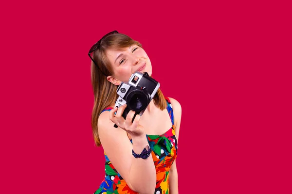 Sorrindo muito jovem mulher loira em vestido multicolorido com óculos de sol na cabeça e uma pulseira na mão segurando a câmera de foto retro. — Fotografia de Stock