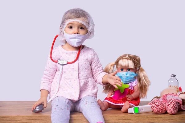 Фото гарної маленької дівчинки, яка грає з лікарем з ляльками в білій текстильній масці для обличчя і тримає стетоскоп на шиї . — стокове фото
