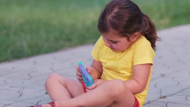 Little cute girl brunetka siedzi na ścieżce w parku w żółtym t-shirt i czerwone szorty gra w pop to gospodarstwa w jej rękach. — Wideo stockowe