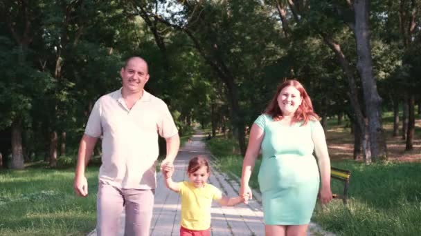 Familia de tres caminando a lo largo del camino en el parque en el fin de semana de verano, ambos padres sostienen a la niña a las manos y saltan juntos. — Vídeo de stock