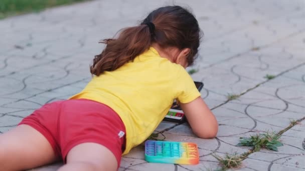 Piccola bella ragazza bruna si sdraia sul suo stomaco sul sentiero nel parco piegando le ginocchia e giocando in smart phone, avendo un giocattolo colorato pop vicino a lei. — Video Stock