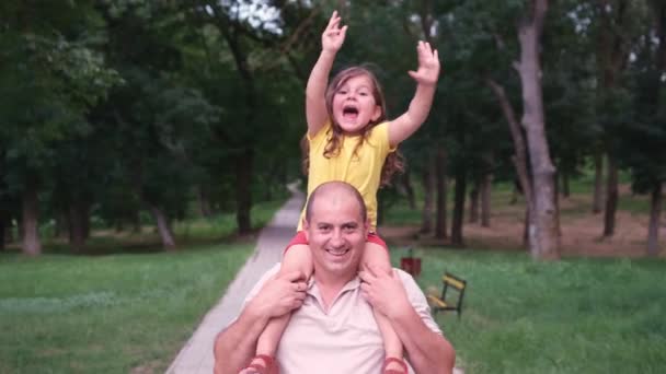 Video frontal ayah dan putrinya bagaimana mereka menghabiskan waktu bersama-sama dengan menjaga putrinya di pundaknya berjalan di taman, meniup ciuman dan mengangkat tangannya melambaikan tangan dengan sukacita. — Stok Video
